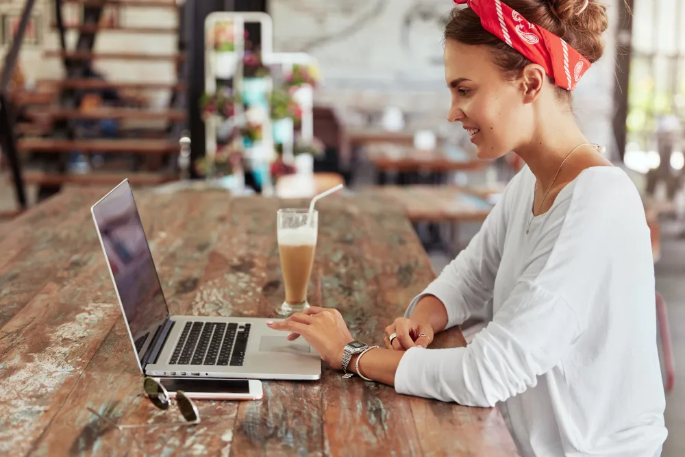 Sideways portrait of smiling female copywriter does remote work on laptop computer, enjoys tasty milk shake at coffee shop, sits alone at wooden table. Woman watches interesting video in website